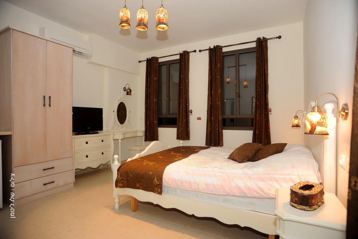 A well-appointed bedroom features a spacious bed covered with a light-colored blanket and accent pillows. Two bedside lamps provide soft lighting, and large windows are adorned with dark curtains. A dresser and a television complete the inviting space.