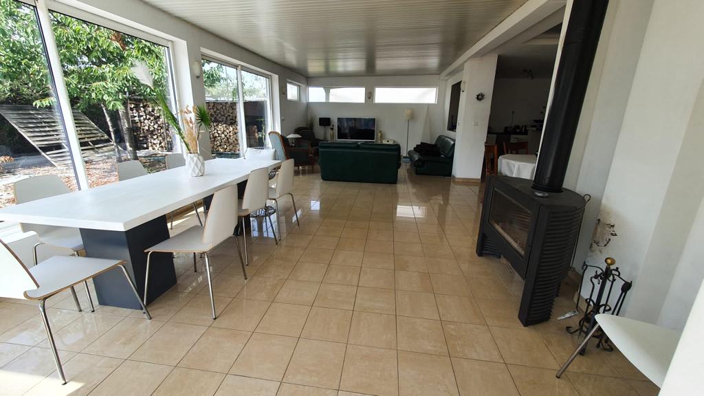 A spacious living area is highlighted by large windows that provide views of the garden. A dining table with chairs sits alongside a cozy seating arrangement, featuring a TV and a fireplace, all set on a polished tiled floor.
