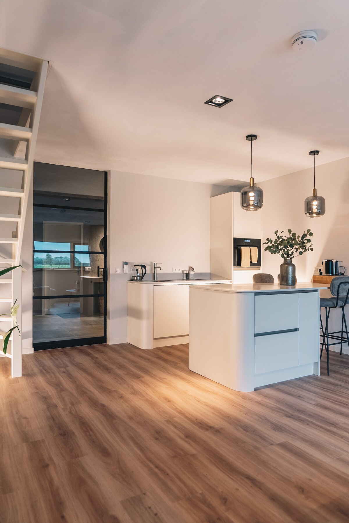 A modern kitchen is seen, featuring a central island with bar stools, sleek cabinetry, and high-end appliances. Natural light floods through large windows, illuminating the wood-like flooring. Stylish pendant lights enhance the contemporary design, creating an inviting space for cooking and dining.