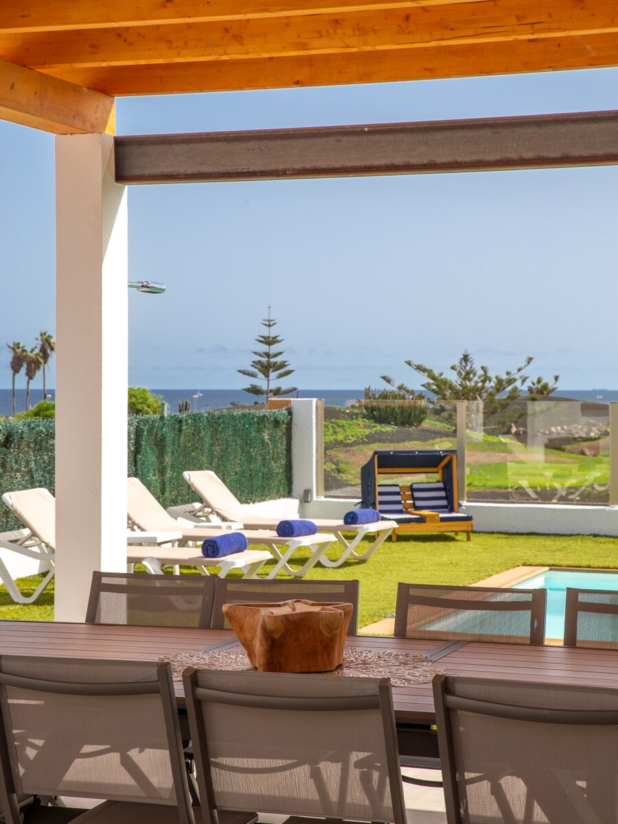 An outdoor dining area features a large table, surrounded by modern chairs. The ocean can be seen in the distance, complemented by loungers and a shaded play area. A vibrant green lawn enhances the space, inviting relaxation and leisure.