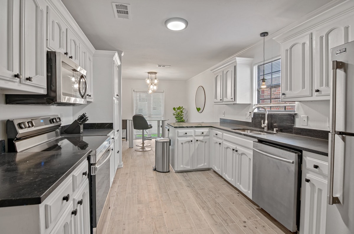 A contemporary kitchen showcases white cabinetry paired with black countertops. Stainless steel appliances are integrated, including an oven and dishwasher. A small dining area is visible through a window, with a round chair and greenery enhancing the space's functionality.