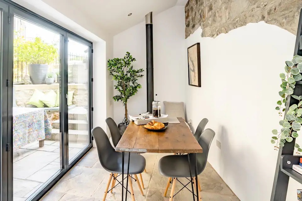 A modern dining area features a wooden table surrounded by sleek grey chairs. Natural light floods through large glass doors, revealing a private courtyard garden beyond. A stone wall adds character, complemented by indoor plants for a touch of greenery.