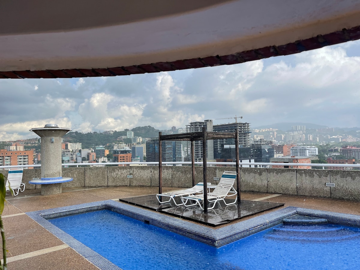 A rooftop area features a refreshing swimming pool with a shallow section. Two lounge chairs are positioned beside the pool, offering a place for relaxation. The scenic view includes a backdrop of urban buildings and hills, with clouds creating a soft ambiance overhead.