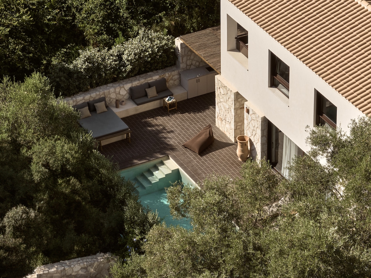 An aerial view of the villa displays a private pool surrounded by lush greenery, with sun loungers positioned for relaxation. The outdoor area is elegantly paved, highlighting the seamless connection between the pool and the villa's architecture.