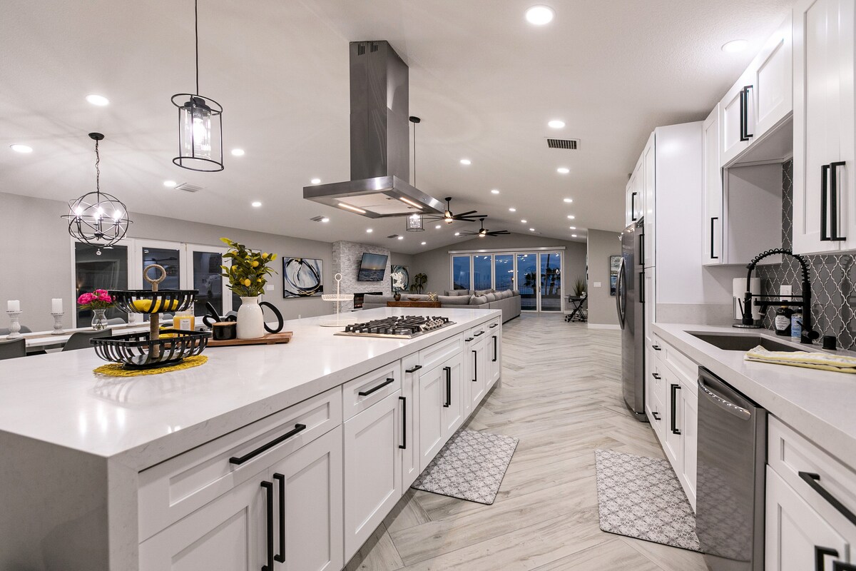 A modern kitchen is highlighted by a spacious quartz island with bar seating. Top-quality appliances are visible, including a stove and refrigerator. Natural light floods the room through large windows, enhancing the sleek cabinetry and decorative accents that create a functional and inviting space.
