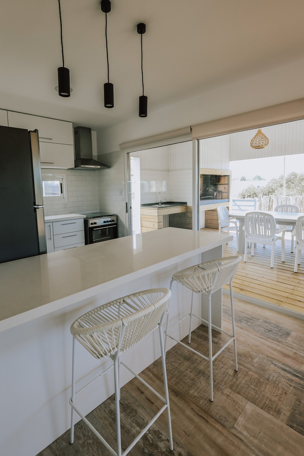 A modern kitchen features a sleek countertop with two high-backed stools. Large windows provide a view of the outdoor deck. The kitchen is equipped with a range, refrigerator, and ample cabinetry, enhancing functionality and natural light within the space.