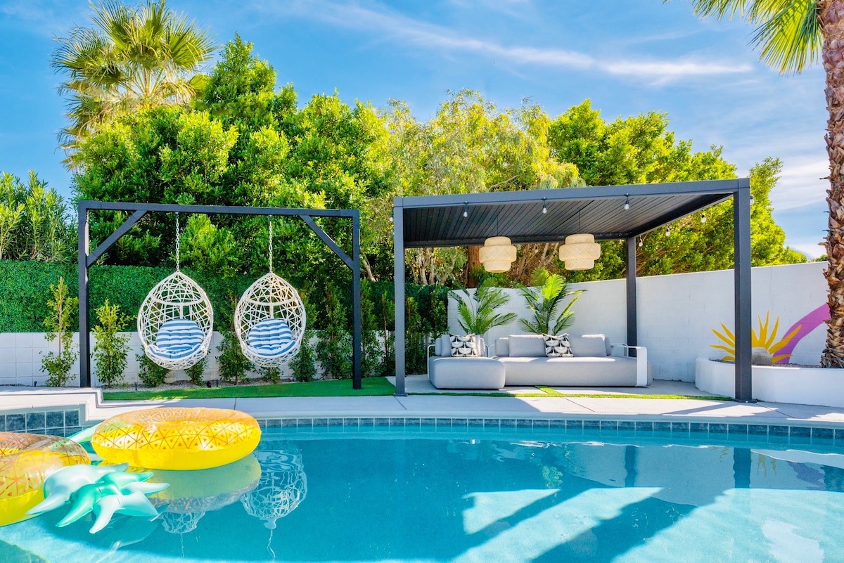 A relaxing outdoor space is highlighted with a shimmering pool surrounded by lush greenery. Two hanging chairs provide seating under a covered area with modern lighting. Brightly colored floatation devices rest by the pool's edge, enhancing the inviting atmosphere for leisurely afternoons.