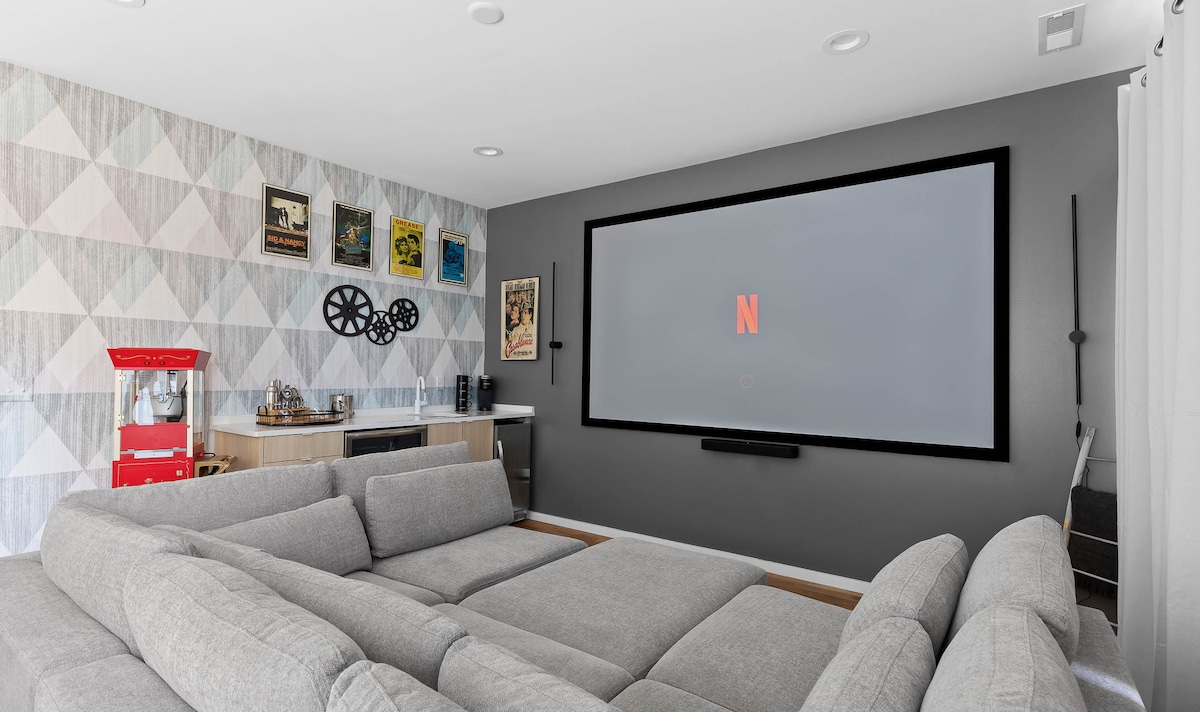 A spacious movie theater room features a large screen displaying the Netflix logo. A cozy sectional sofa in a soft gray color surrounds the space, complemented by a retro popcorn machine and tasteful artwork on the walls.