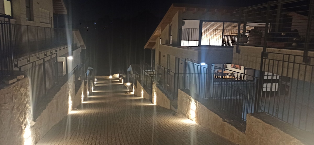 A well-lit pathway is lined with subtle stone walls and illuminated by soft lighting along the edges. The path leads towards a cluster of modern buildings, showcasing wooden roofs and open balconies, creating an inviting atmosphere for evening strolls.