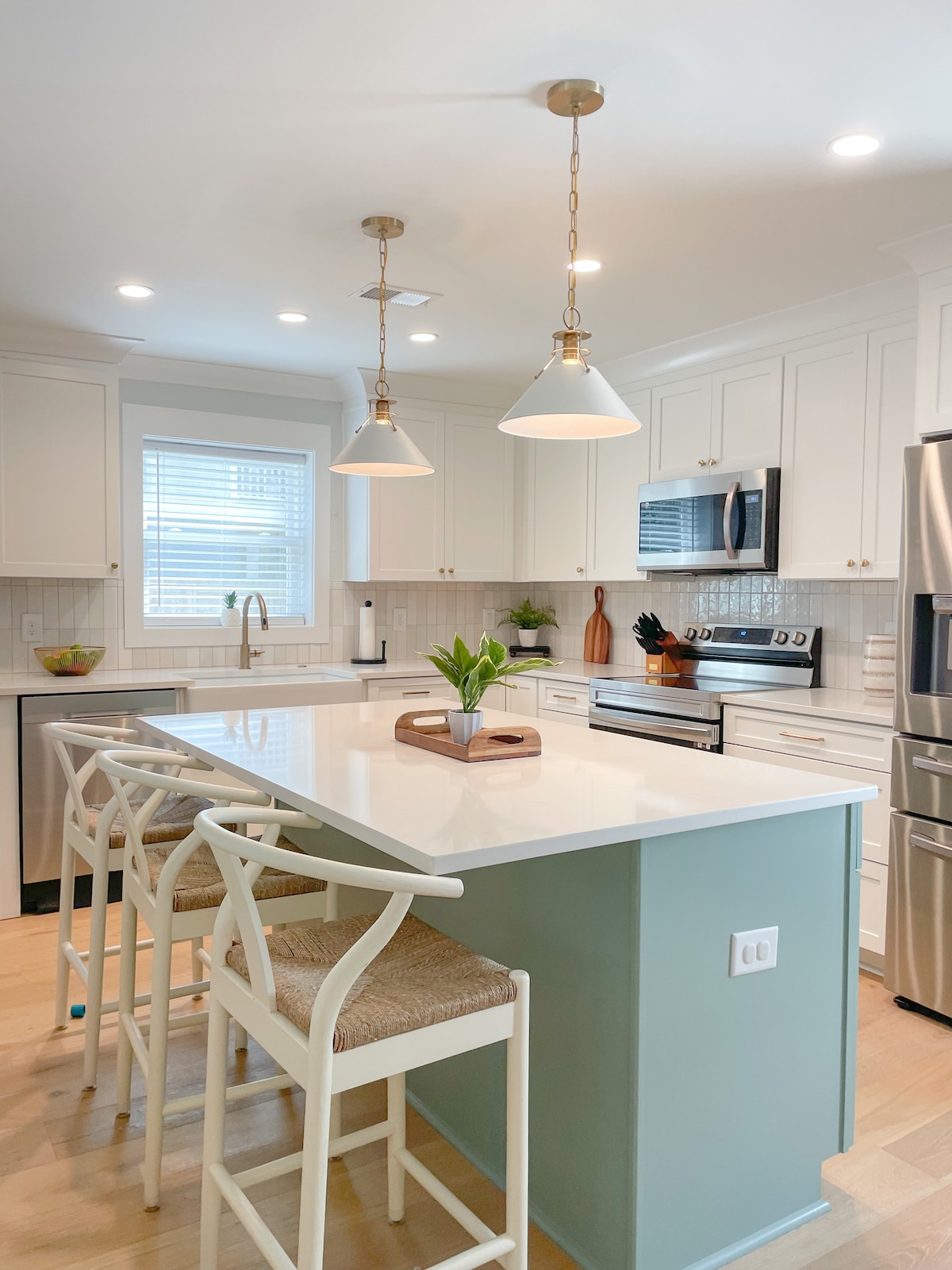 A modern kitchen features white cabinetry and stainless steel appliances. A large island is positioned at the center, accompanied by light-colored seating. Pendant lights hang above, illuminating the space, while a window allows natural light to enter, enhancing the minimalist and functional design.