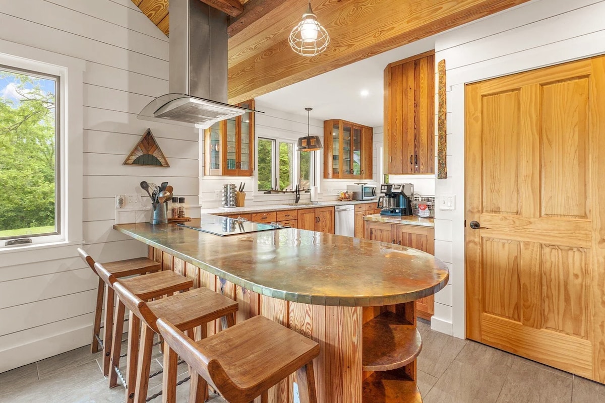 A spacious kitchen is featured, showcasing wood cabinetry and a large island with a unique stone countertop. Four wooden stools are positioned along the island. Natural light enters through several windows, highlighting the room's warm, rustic design.