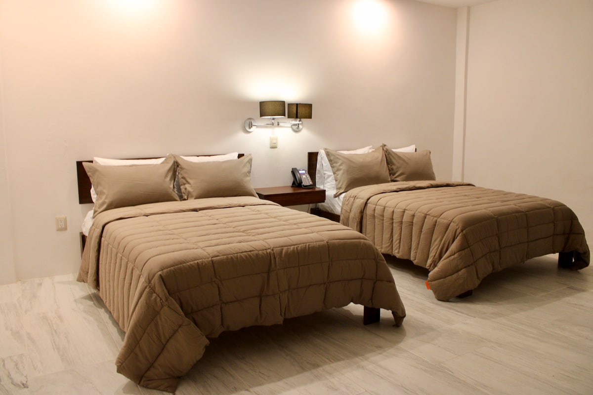 The guest room features two spacious beds, each dressed in neutral-toned bedding. A simple nightstand is positioned between the beds, topped with modern wall sconces that provide soft lighting. Light-colored floors enhance the room's bright and clean aesthetic.