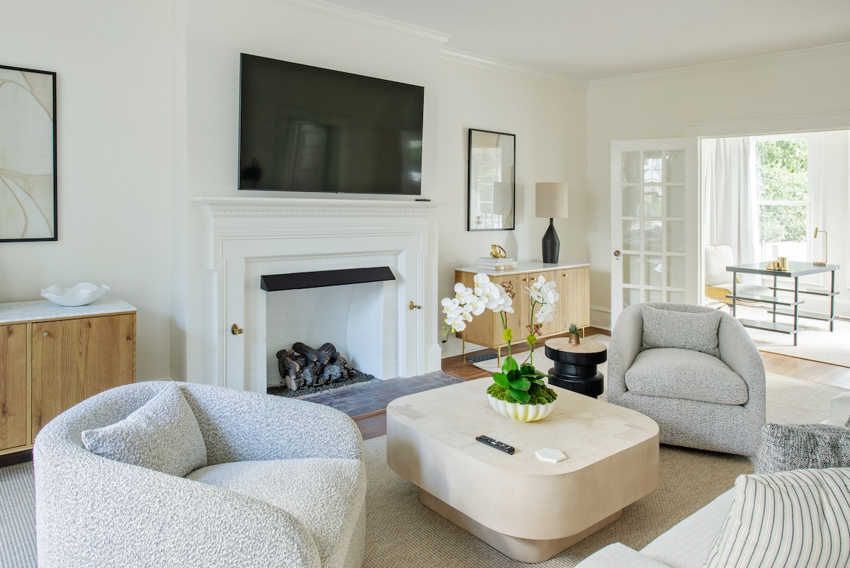 A spacious living area presents a cozy arrangement with two rounded armchairs and a central coffee table. A large flat-screen TV is mounted above a decorative fireplace, surrounded by minimalistic décor. Natural light filters in through French doors leading to an outdoor space.