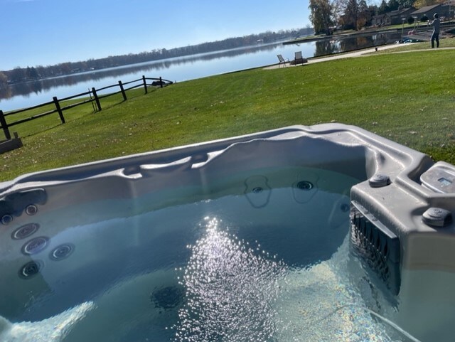 A hot tub is situated on a well-maintained lawn, offering serene views of the lake. The water reflects sunlight, creating sparkling patterns. In the background, a wooden fence and comfortable lounge chairs enhance the peaceful, outdoor setting by the waterfront.