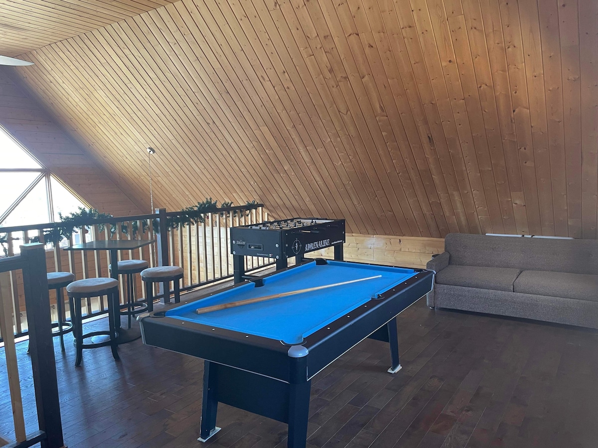 An inviting game room features a blue pool table with a cue stick resting on it. The wooden walls add warmth, complemented by light streaming through large windows. Two bar stools are positioned nearby, alongside a comfortable couch for relaxation.