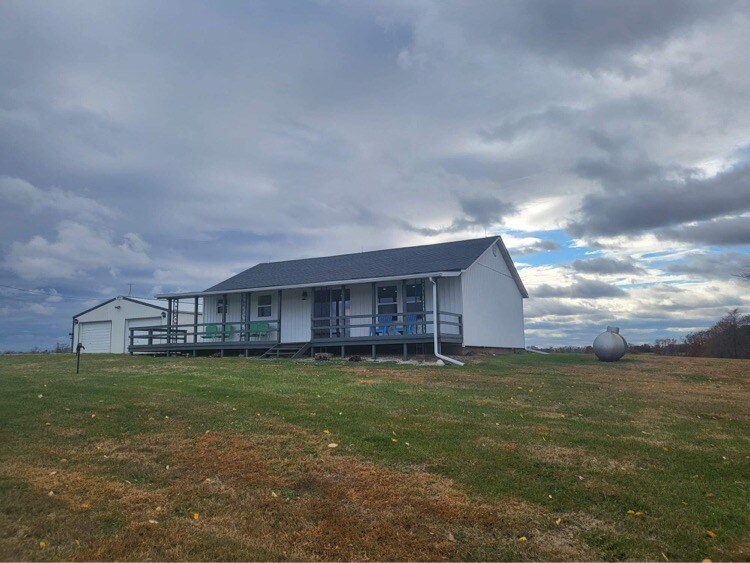 The exterior of the remodeled farmhouse is showcased against a cloudy sky. A spacious lawn surrounds the home, and a covered porch with blue chairs is visible. A storage shed and a round propane tank are positioned nearby, adding to the rural charm.