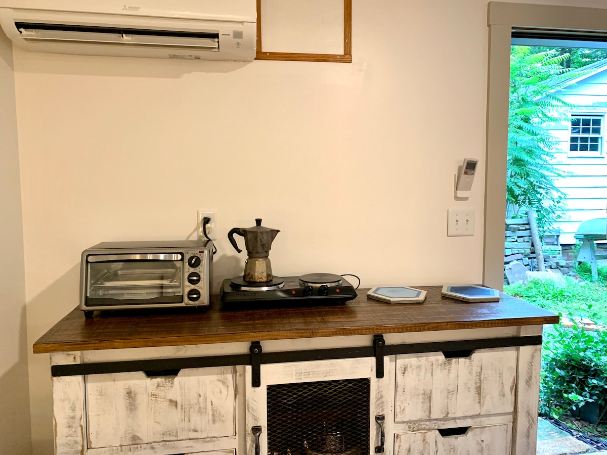 A kitchenette area is displayed featuring a small electric kettle, a toaster oven, and a two-burner hot plate on a wooden countertop. White cabinetry with a rustic finish adds character. Natural light enters through a window, providing a glimpse of the greenery outside.
