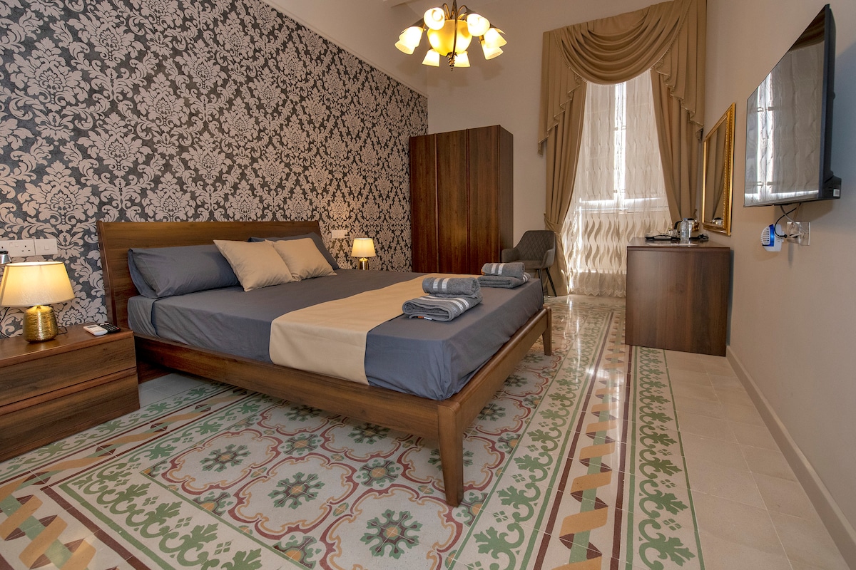A spacious room features a large bed with neatly arranged linens and towels. Traditional Maltese tiles cover the floor, complemented by a patterned accent wall. Natural light enters through a large window, which is draped with elegant curtains. Additional furniture includes a wardrobe and a bedside table.