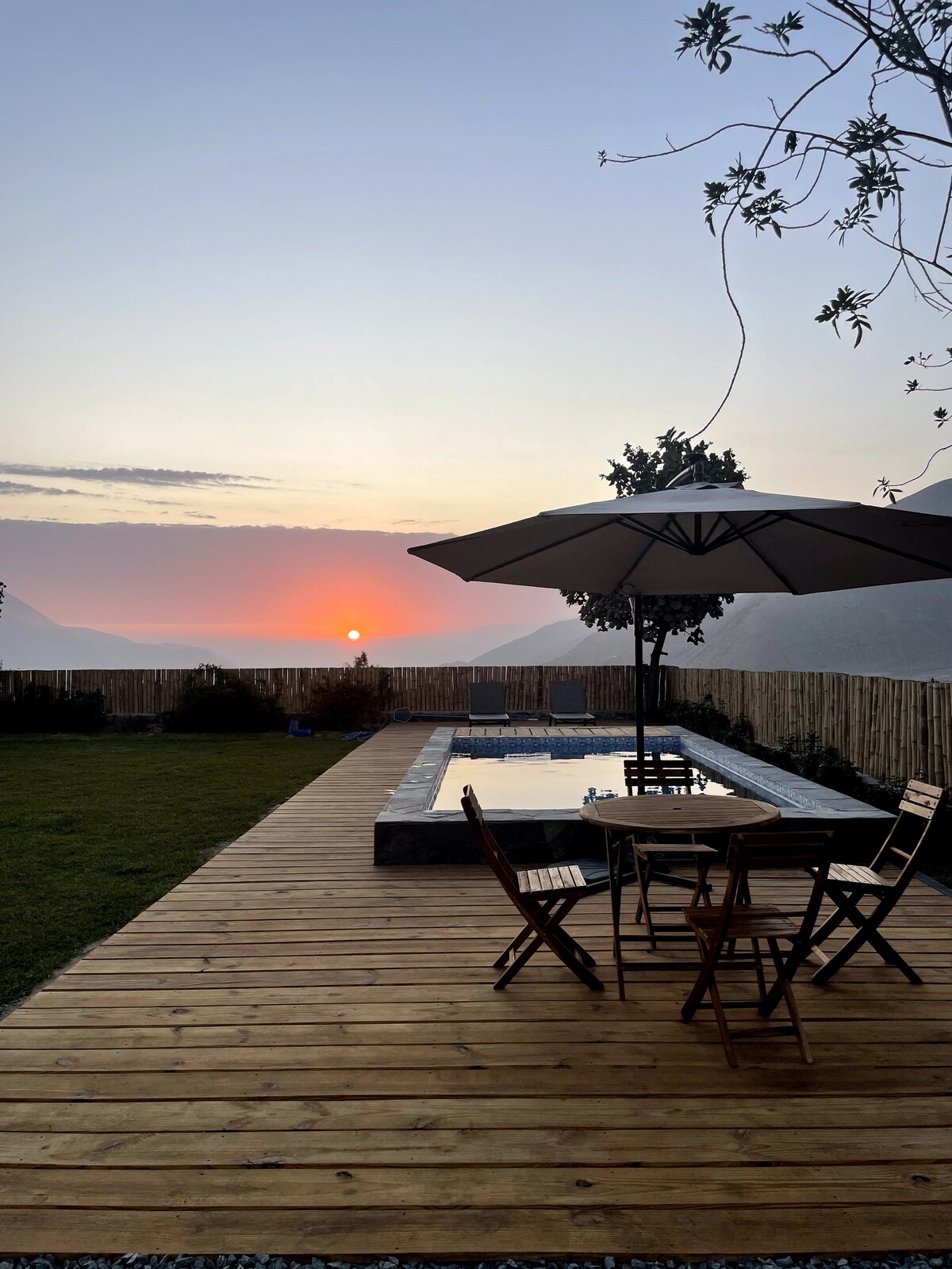 A sun setting beyond rolling hills is viewed from a wooden deck. A swimming pool is situated nearby, complemented by a round table and four folding chairs under a large umbrella, providing a serene outdoor space for relaxation.