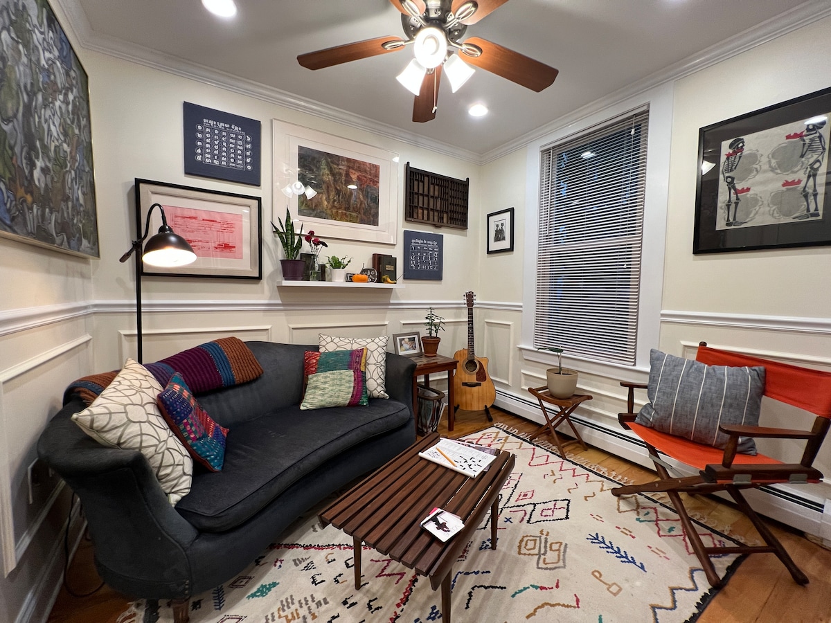 A cozy seating area is presented with a dark sofa adorned with colorful cushions. An acoustic guitar leans against the wall, while a wooden coffee table sits in the center. The walls are decorated with framed artworks, and light from a ceiling fan illuminates the space.