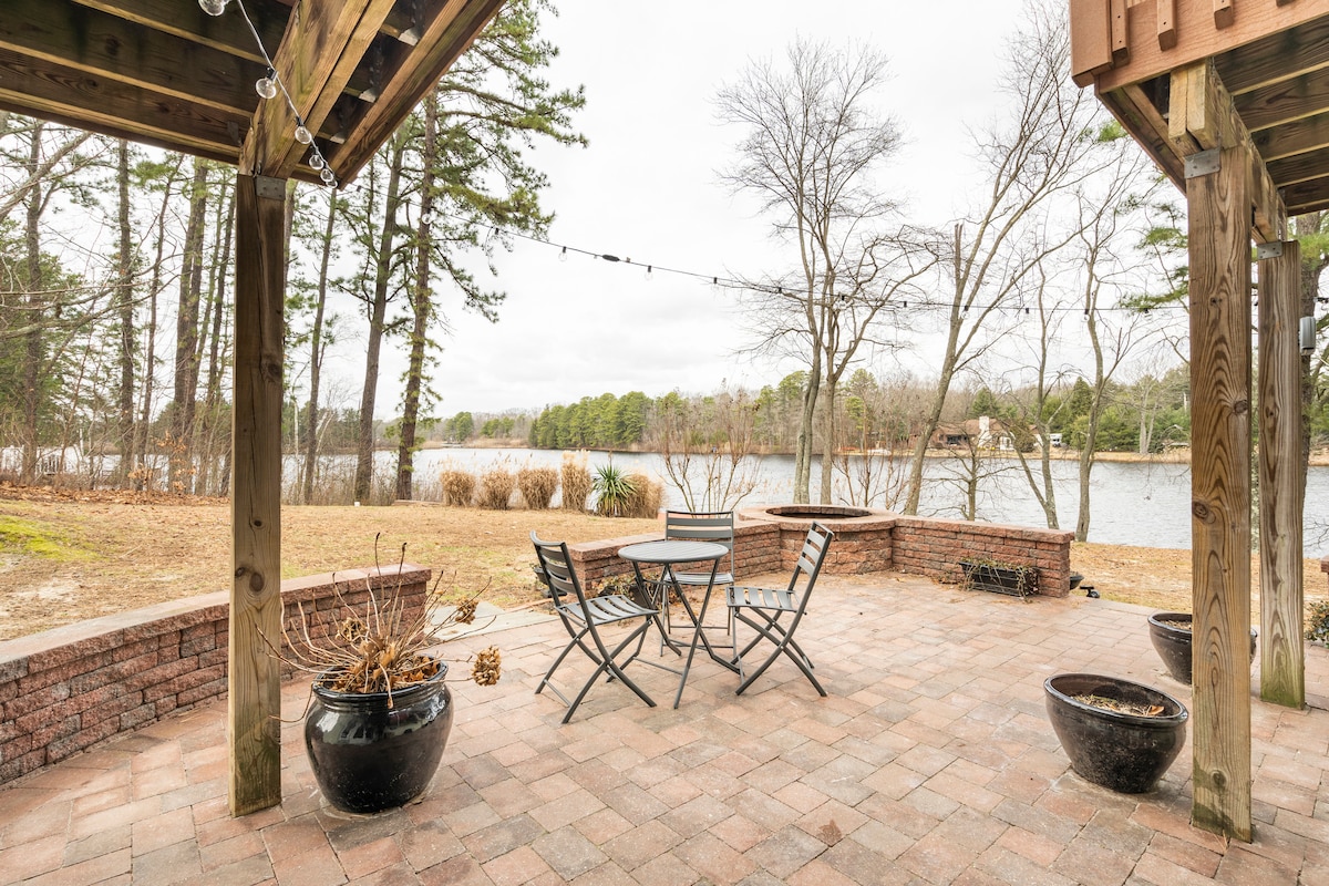 An outdoor patio area features a stone-paved floor, surrounded by wooden beams and overhead string lights. A table with four chairs faces a serene view of the lake and surrounding trees in the distance. Potted plants add greenery to the space.