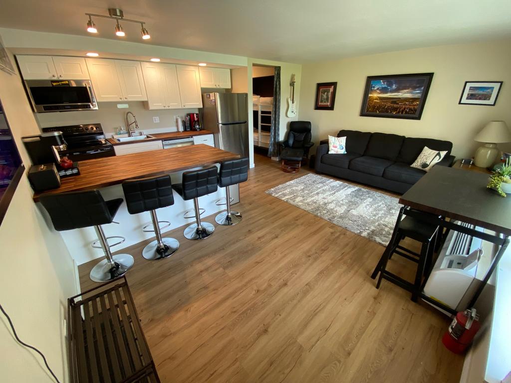 A well-appointed living area reveals a stylish kitchen with modern appliances and a breakfast bar featuring four stools. A comfortable dark sofa faces a rug-covered floor, while artwork adorns the walls. Natural light brightens the space through multiple windows, enhancing the overall ambiance.