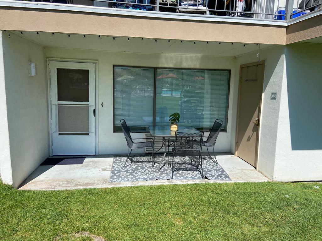 A private patio area features a glass table surrounded by four chairs, situated next to a large window. A door leads inside, while the green lawn provides a pleasant setting. Sunlight illuminates the space, creating a relaxed outdoor atmosphere.