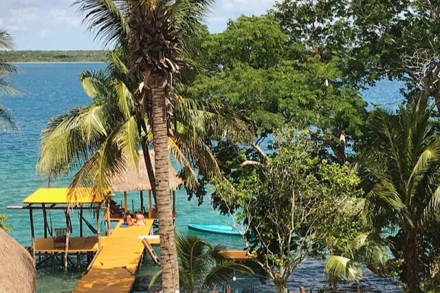 A wooden dock extends into the clear waters of the lagoon, surrounded by lush palm trees and tropical foliage. A covered palapa sits at the edge, providing shaded space for relaxation, with a small boat visible near the shore.
