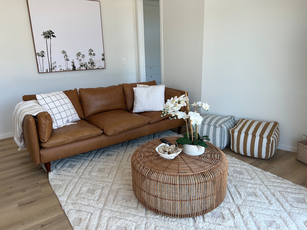 A cozy living area is furnished with a leather sofa and decorative cushions. A round woven table stands at the center, adorned with a potted plant and decorative items. A textured rug complements the space, while a wall art piece adds a subtle touch of elegance.