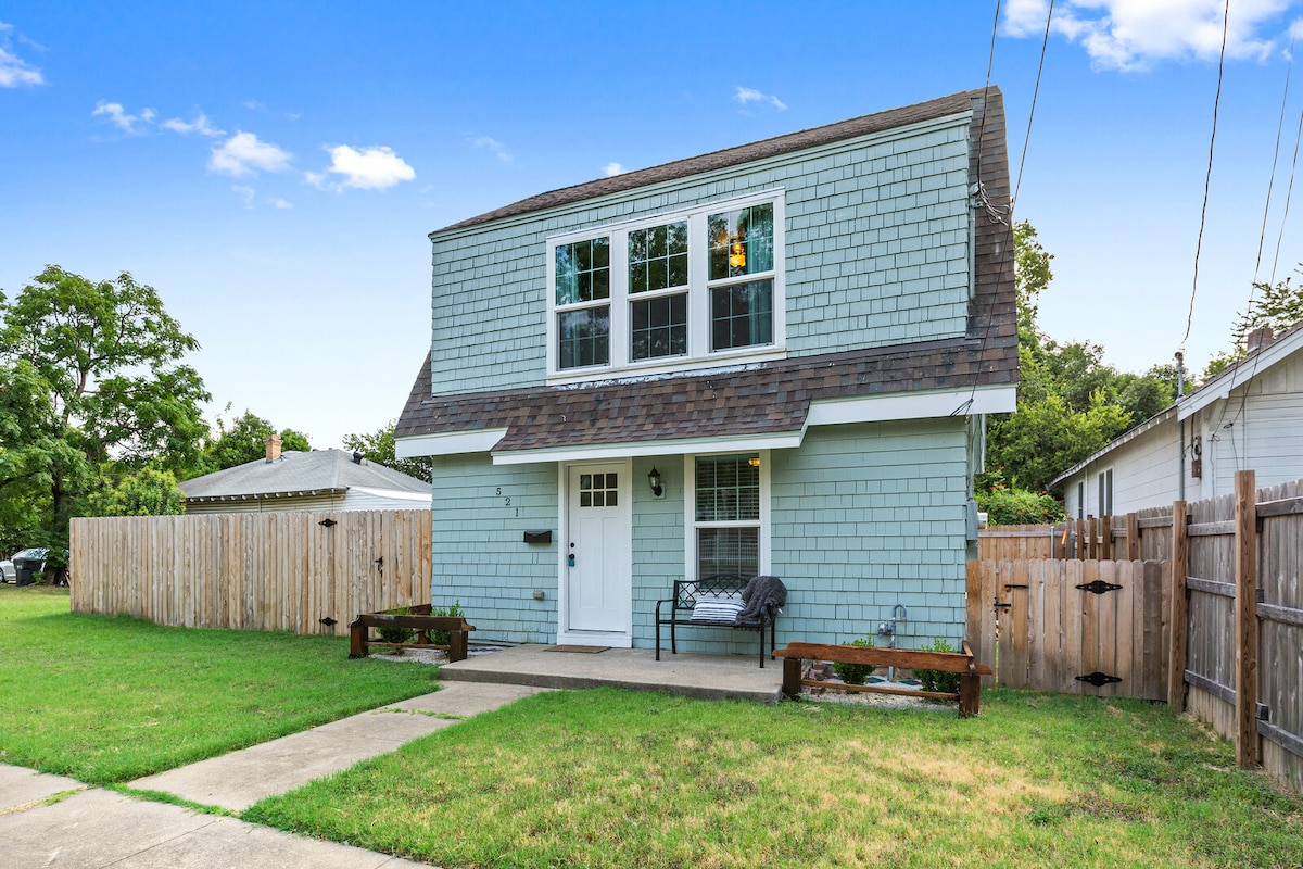 A two-story home is presented, featuring a light blue exterior with multiple windows. A welcoming entrance is highlighted by a small covered porch with seating. The surrounding area includes a grassy lawn and wooden fences lining the property.