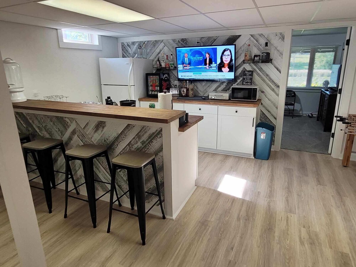 A modern kitchen space features a bar with four high stools and a wooden countertop. A large white refrigerator and various appliances are visible, while a smart TV displays a news channel. Natural light floods in through nearby windows, illuminating the light-colored flooring.