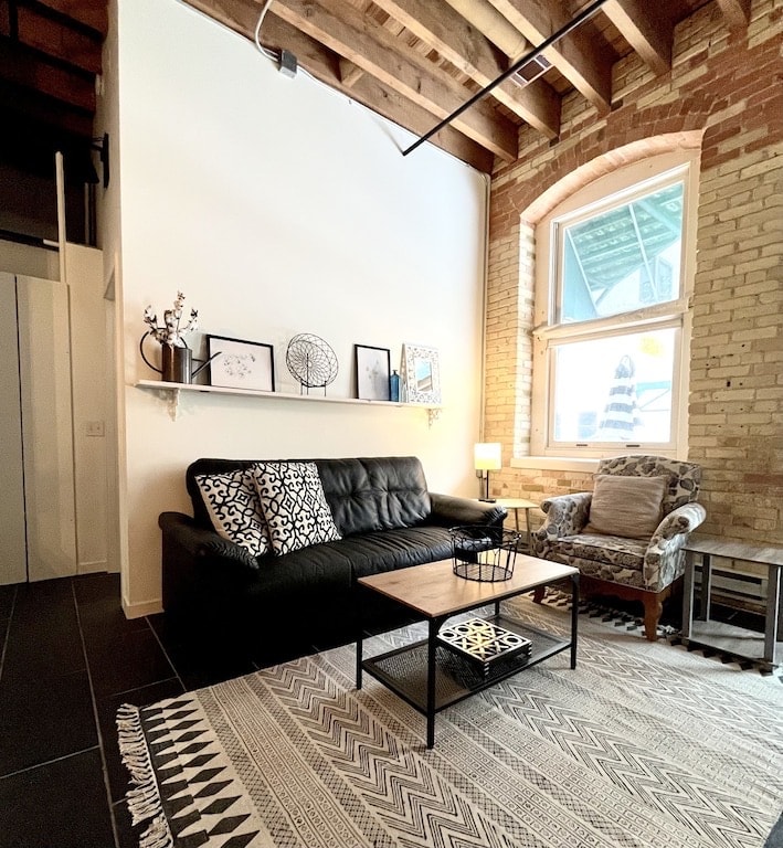 A cozy living area is showcased with a black sofa and a patterned armchair beside a wooden coffee table. Natural light filters through the arched window, illuminating the shelves with framed wall art. A textured area rug adds warmth to the space.