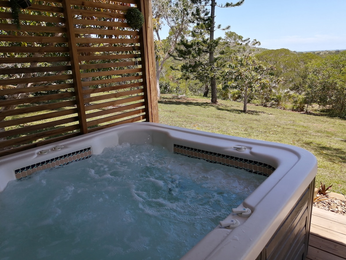 A private hot tub is located on a wooden deck, surrounded by slatted wood panels. The bubbling water reflects the natural light, while views of a green landscape and trees are visible in the background.