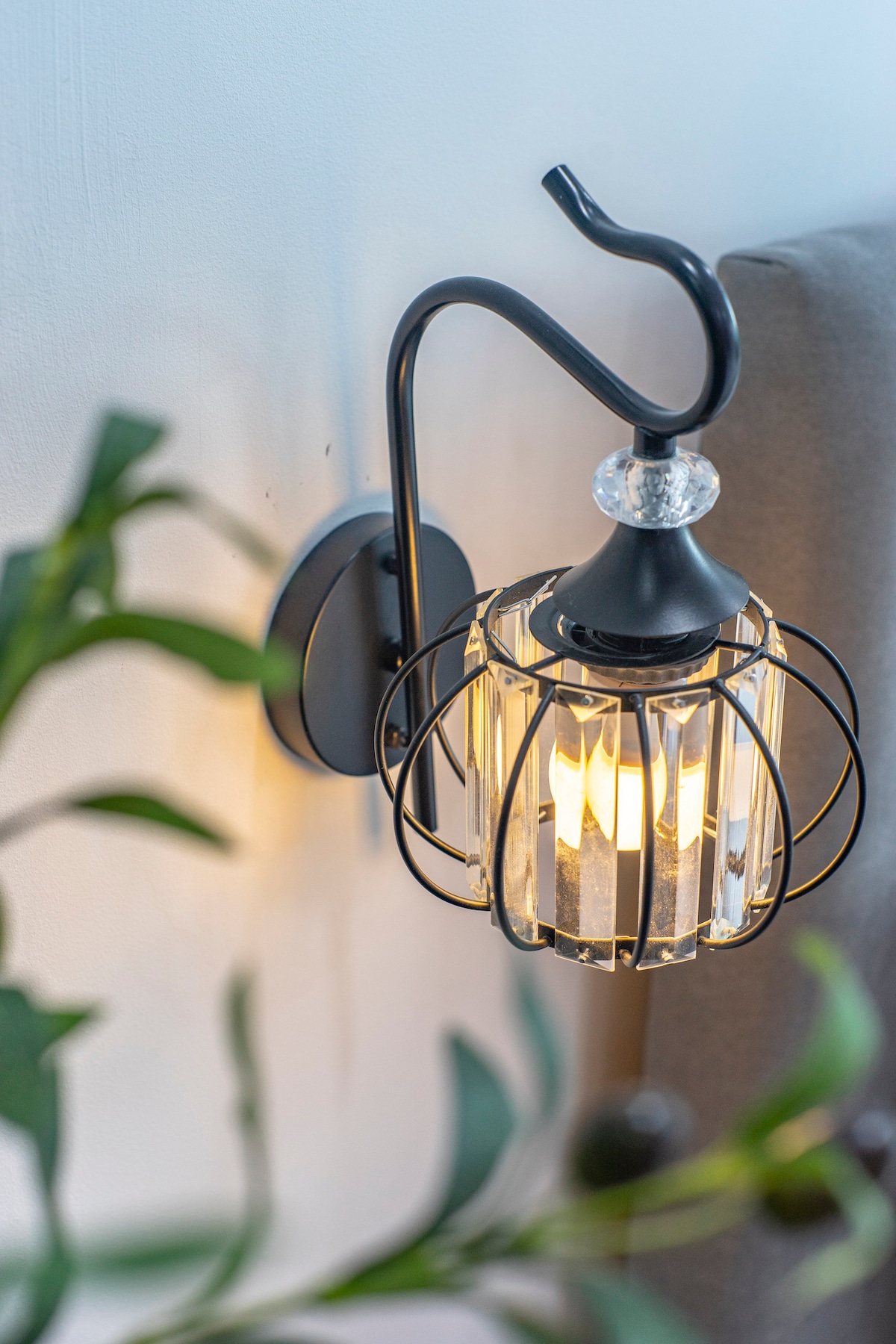 A stylish wall sconce is mounted beside a bed, featuring a black metal frame with a crystal enclosure. The soft golden light illuminates the surrounding area, contrasting with the greenery from nearby plants.