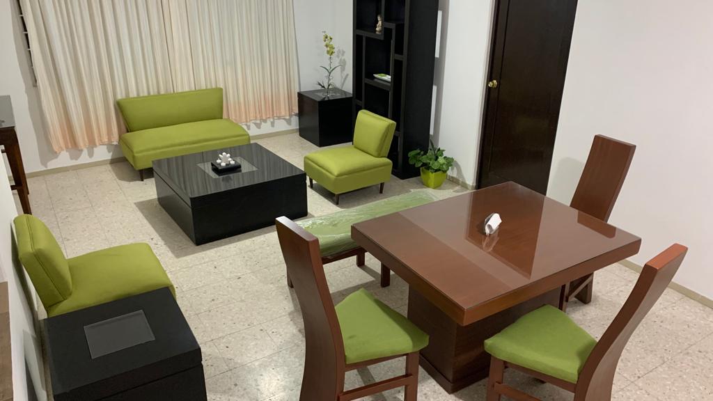 A cozy living area features a dark coffee table at its center, surrounded by two green armchairs and a green sofa. A wooden dining table is positioned nearby, accompanied by four matching chairs. Natural light filters through sheer curtains, highlighting the neutral-toned walls.
