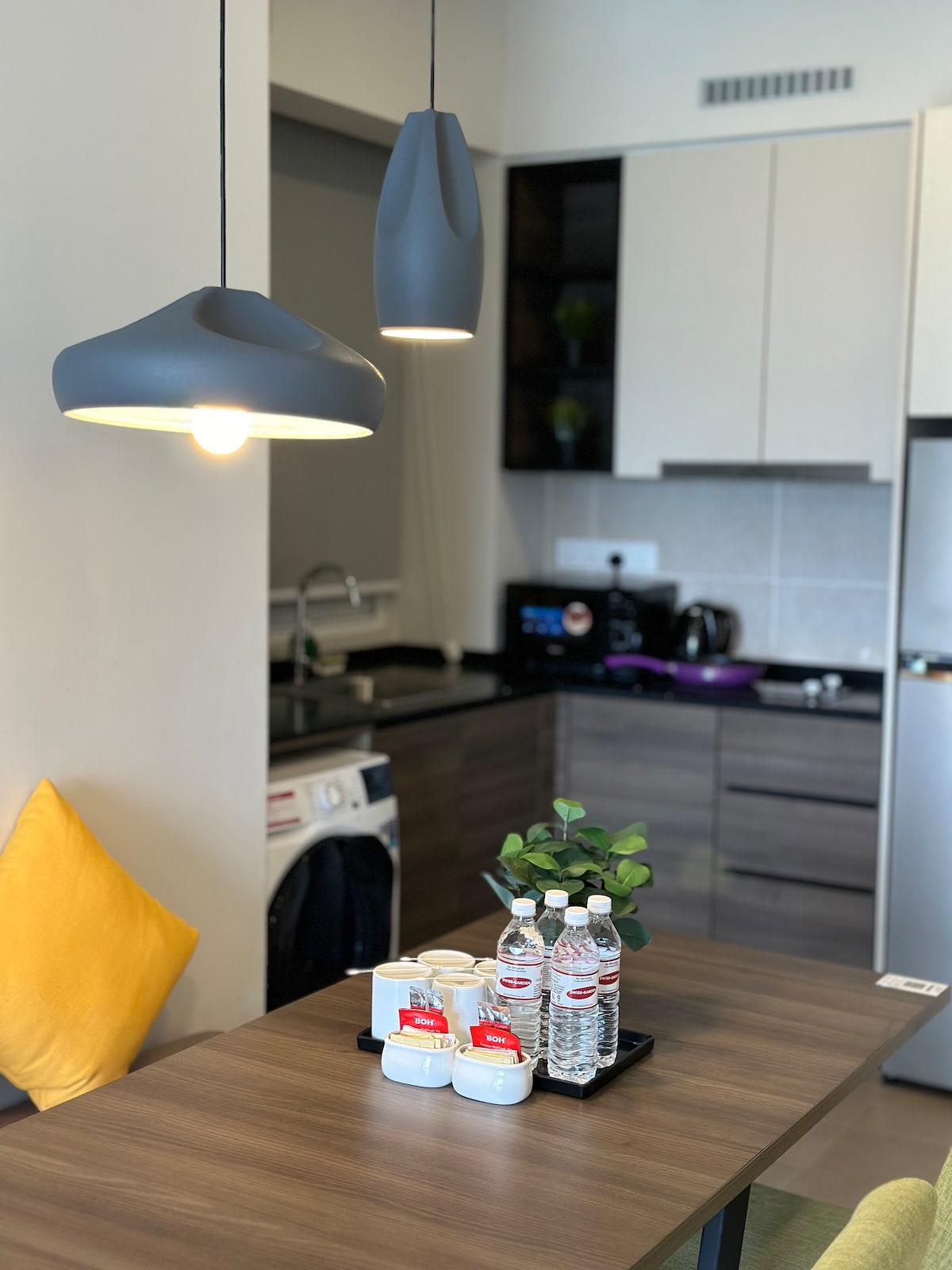 A modern kitchen space is highlighted with sleek cabinetry and contemporary appliances. A dining table is set with a small plant and bottled water, accompanied by snack containers. Two pendant lights add gentle illumination above the table, while accents of yellow provide a pop of color.
