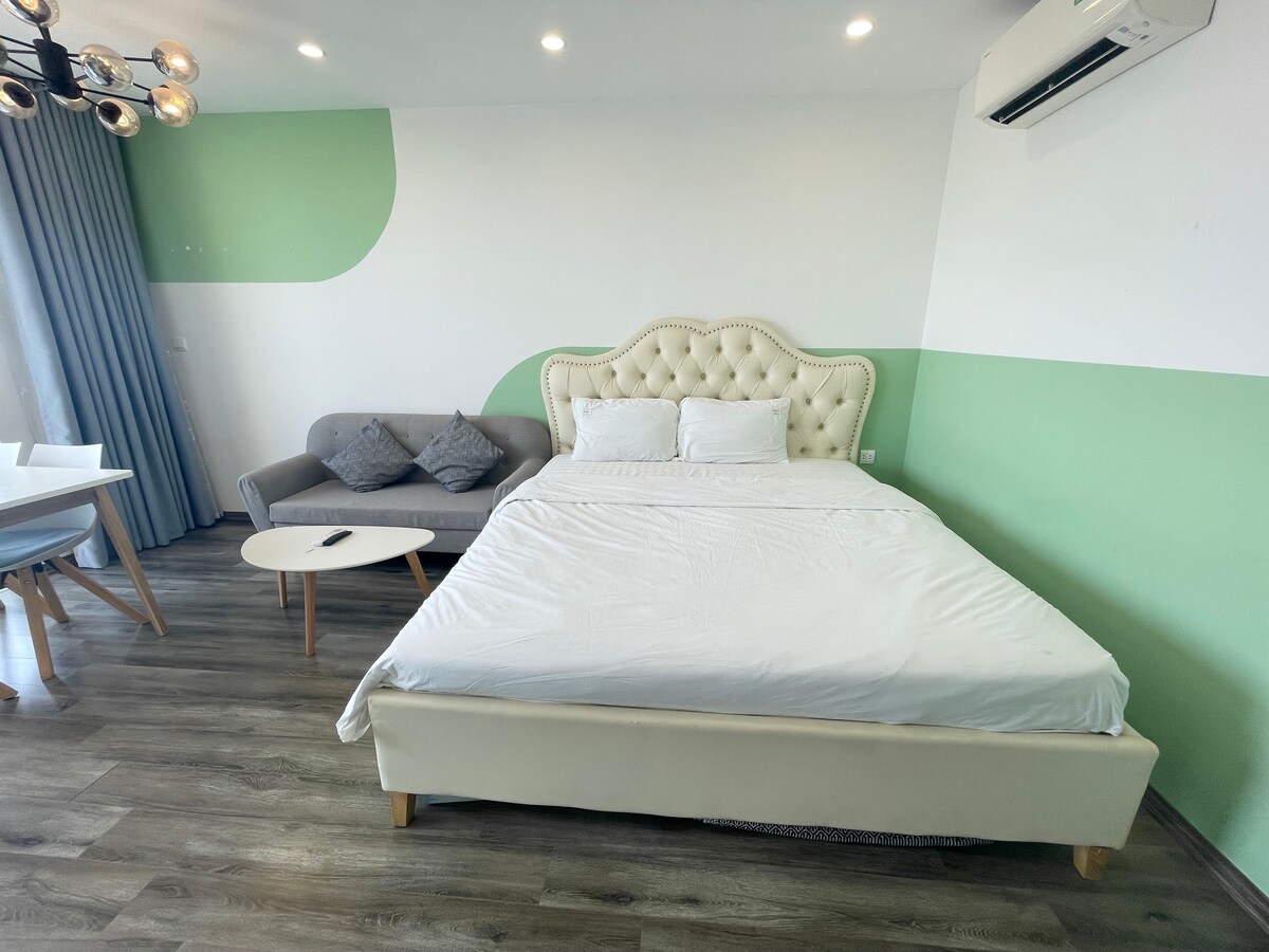 A stylish bedroom features a spacious bed with a tufted headboard and crisp white linens. A comfortable sofa sits against a light green accent wall, complemented by streamlined furniture. Natural light enters through sheer curtains, enhancing the serene atmosphere.