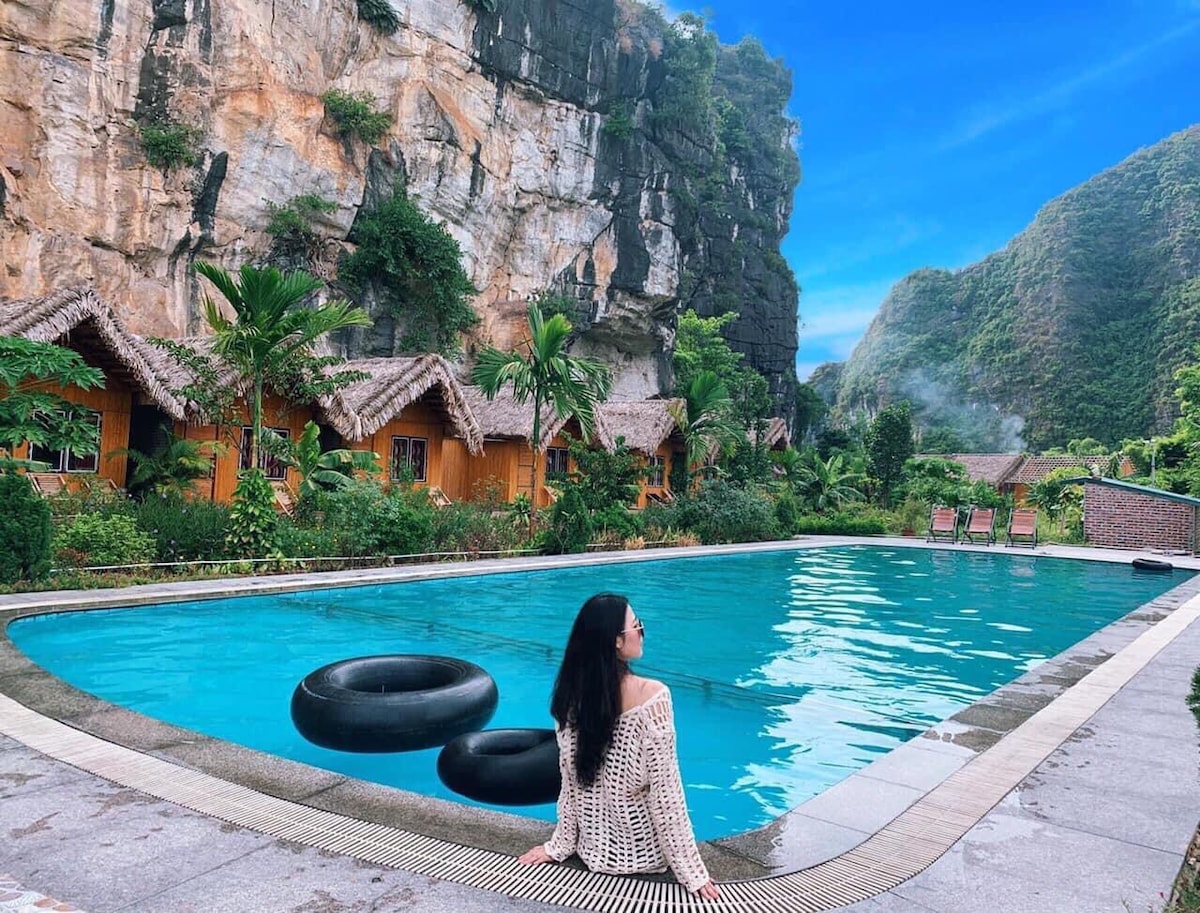 A tranquil swimming pool features clear turquoise waters, complemented by natural stone surroundings and lush greenery. Two circular inflatable rings rest at the pool's edge. Traditional-style accommodations with thatched roofs are visible in the background, nestled against a striking rocky cliff.
