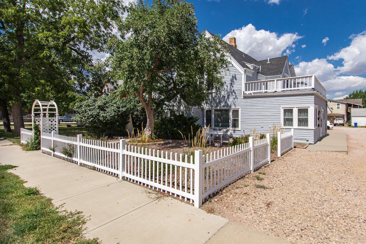 The exterior of a charming residence is displayed, featuring a light blue façade and a white picket fence. A gravel area is visible in front, along with landscaped shrubbery. A mature tree provides shade, while a balcony can be seen on the upper level under a partly cloudy sky.