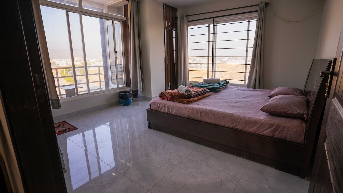 A spacious bedroom features a large bed with neatly arranged linens. Natural light streams through large windows, providing a view of the surroundings. A couple of towels are placed on the bed, and a wastebasket is positioned near the window. The floor is tiled and well-maintained.