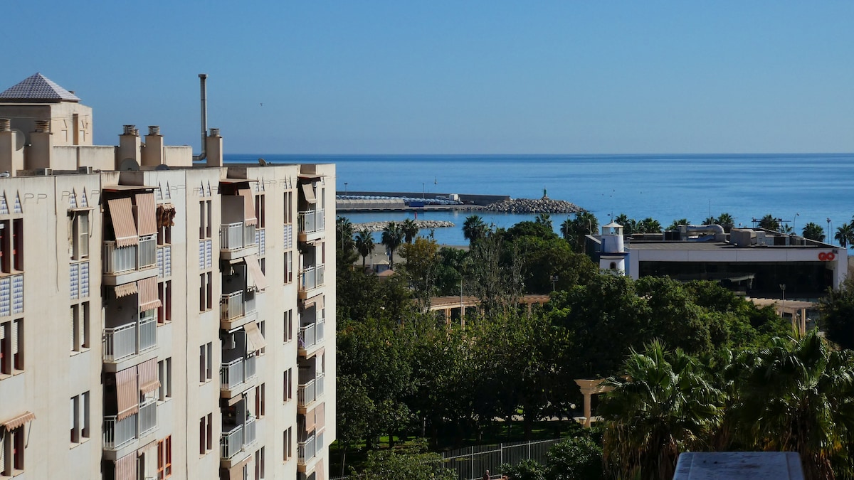 A view reveals a colorful coastal scene, featuring clear blue waters and a distant jetty. Lush greenery surrounds a well-maintained park visible below, while modern buildings create a backdrop along the shoreline.