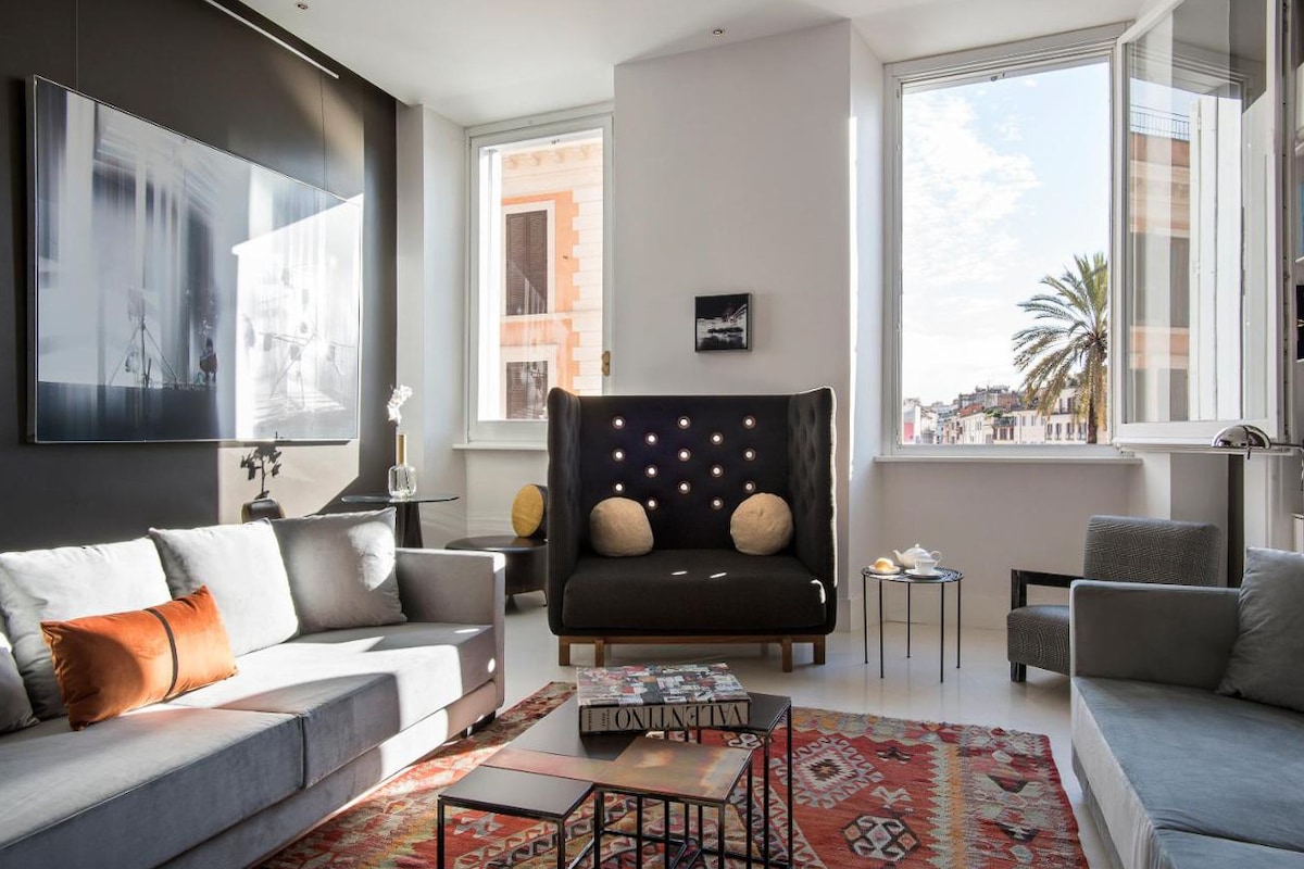 A bright living area is characterized by large windows allowing natural light to flood the space. Stylish seating includes a gray sofa and a unique black couch with decorative accents. A coffee table rests on a patterned rug, complemented by a small side table and decorative artwork.