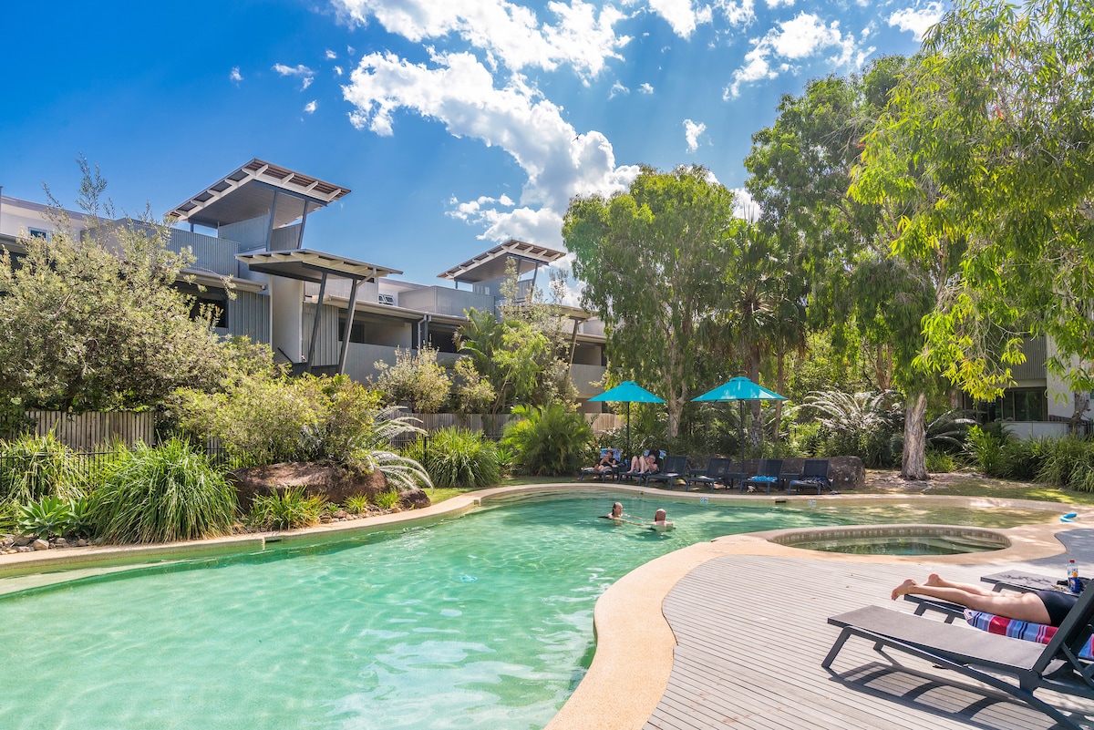 A serene heated pool is surrounded by lush greenery and stylish lounge chairs. Umbrellas provide shade for relaxation. The pool's gentle curves intersect with wooden decking, allowing for easy access, while the buildings in the background complement the natural landscape.