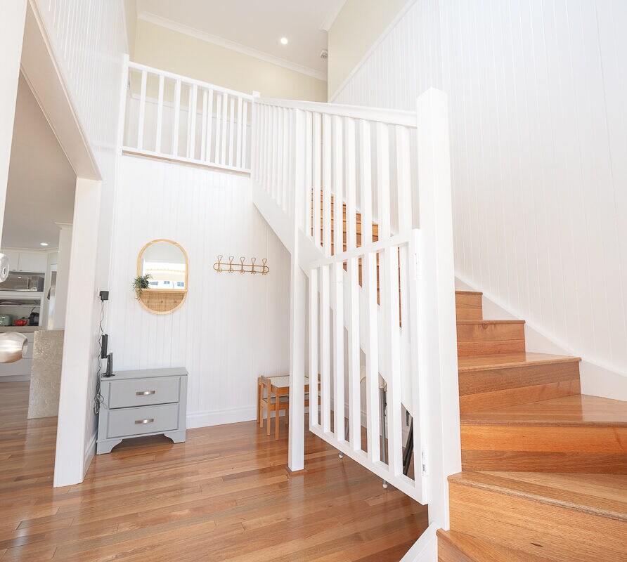 A bright entryway features wooden stairs leading to the upper level. A circular mirror is mounted on the wall beside a small console table. The light-coloured walls contribute to the airy ambiance, with polished wooden flooring covering the space.