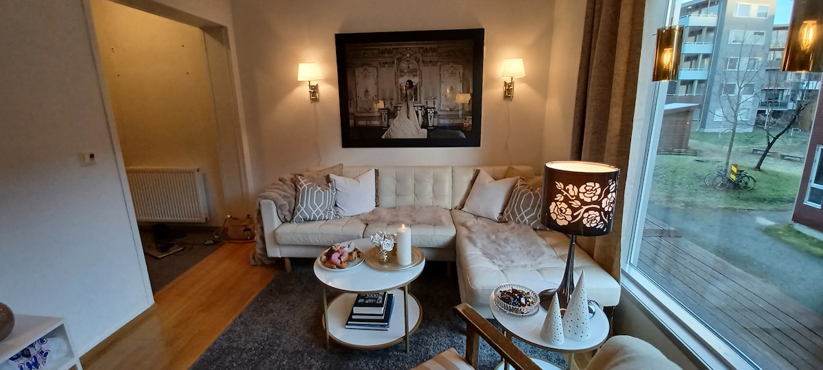 A cozy living area features a light-colored sectional sofa paired with a round coffee table. Ambient lighting is provided by wall sconces and a decorative lamp. A large window reveals a glimpse of the outdoor area, enhancing the space's openness.