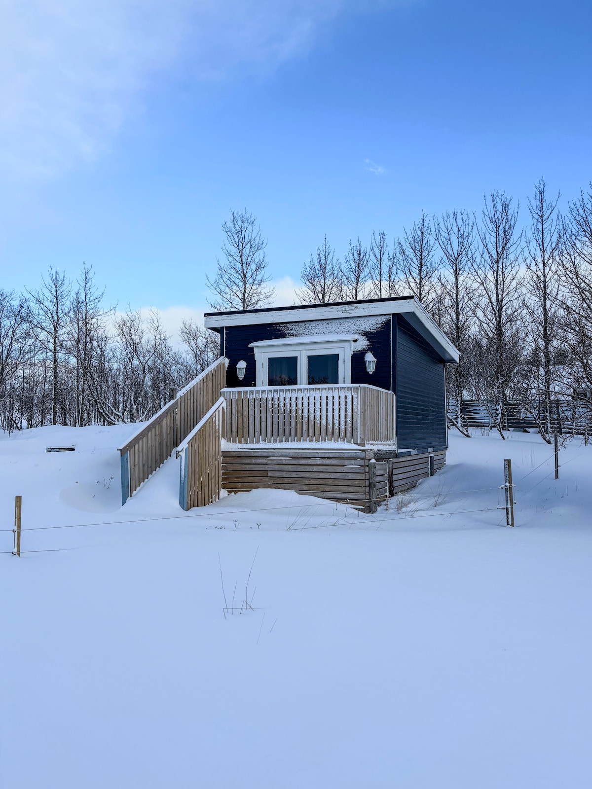 A charming cottage is set against a backdrop of snow-covered ground and sparse trees. The dark exterior is complemented by a light-colored porch and white snow adorning the stairs leading up to the entrance, inviting exploration.
