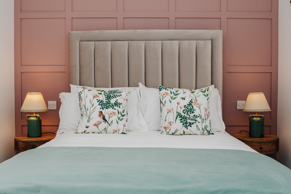 The bedroom features a plush headboard with a subtle pink panelled wall behind it. A spacious bed is adorned with elegant bedding and decorative pillows, complemented by bedside tables and soft lamps on either side, creating a tranquil and inviting space.
