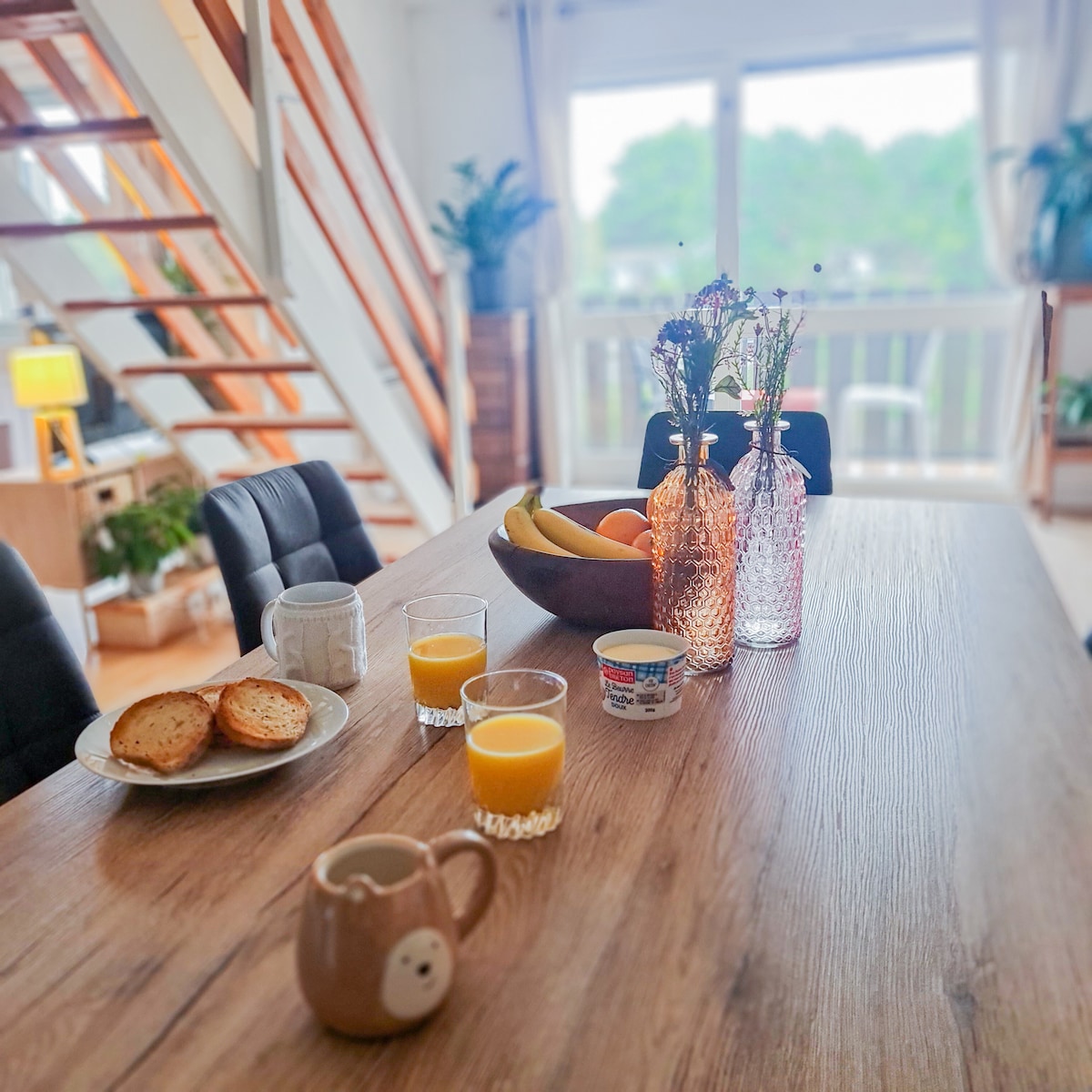 A dining table is set with plates of bread and yogurt, accompanied by glasses of orange juice. Two decorative glass bottles hold fresh flowers, while a bowl of bananas rests at the center. A warm ambiance is created by natural light filtering through a nearby window.