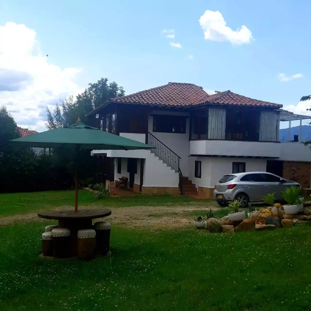 A two-story home with a tiled roof is set amidst a grassy area. A round table with seating is shaded by a large umbrella. A vehicle is parked nearby, with natural greenery framing the scene and a clear sky above, enhancing the tranquil environment.