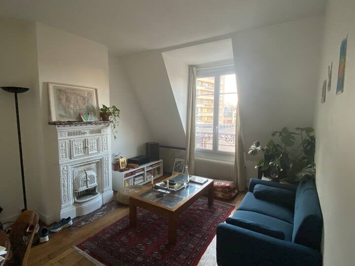 A bright living room features a blue sofa positioned beside a coffee table, surrounded by greenery. A decorative fireplace adds character to the space. Sunlight filters through a large window, revealing views of the neighborhood while illuminating the light-colored walls.